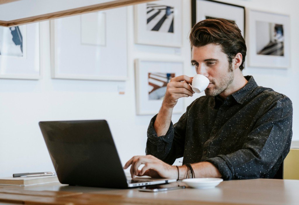 A professional man sitting at a wooden desk, focused on his laptop while drinking coffee in a modern office.